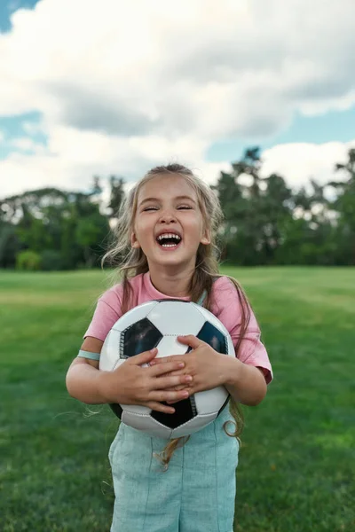 Retrato de niña alegre sosteniendo el fútbol y riéndose de la cámara mientras juega con sus padres en el parque — Foto de Stock