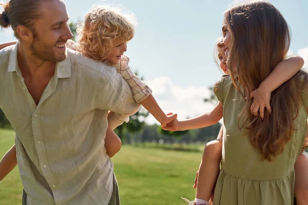 Små barn, pojke och flicka håller varandra i händerna medan de njuter av piggyback rida på baksidan av sin mamma och pappa. Familjevandring i grön sommarpark — Stockfoto