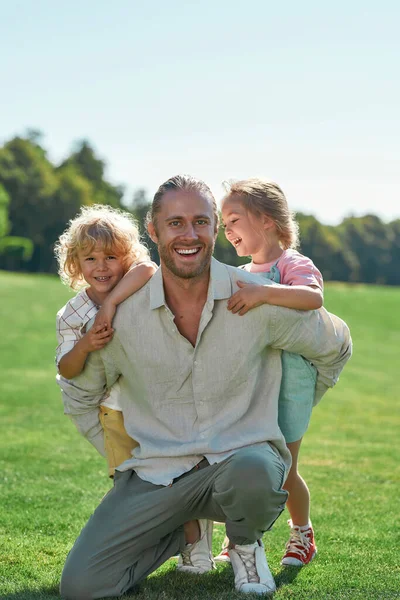 Alegre papá sonriendo a la cámara, posando junto con dos lindos niños pequeños, hija e hijo en el campo de hierba en el parque de verano —  Fotos de Stock