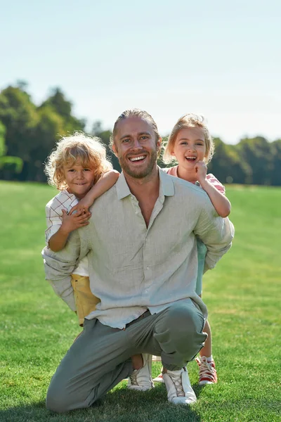 Mosolygó apa a kamerába néz, pózol együtt két aranyos kisgyerekkel, lányával és fiával a fűpályán a nyári parkban. — Stock Fotó