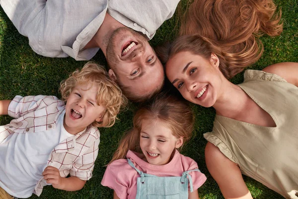 Vista superior de los padres jóvenes felices con su hija pequeña y su hijo tumbado en la hierba verde en el parque de verano —  Fotos de Stock