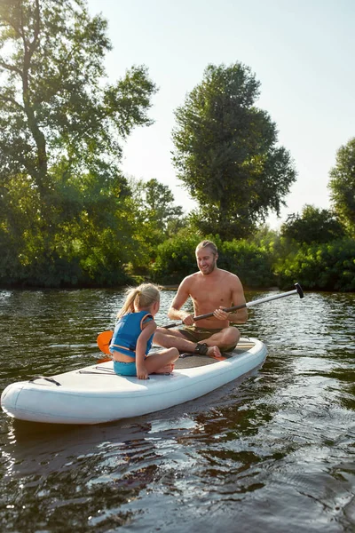 Vater bringt seiner kleinen Tochter bei, wie man paddelt, während man auf einem Fluss surft, der an einem Sommertag von der schönen Natur umgeben ist — Stockfoto