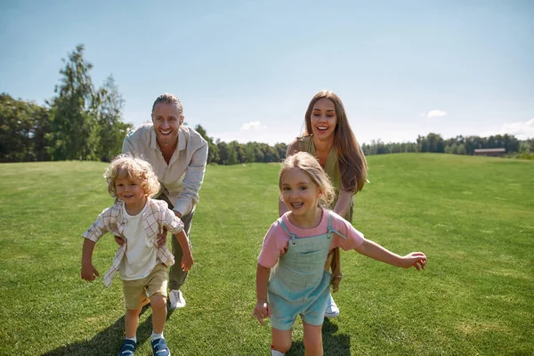 Glada unga föräldrar leker fånga tillsammans med två små barn, pojke och flicka i grön park på en sommardag — Stockfoto