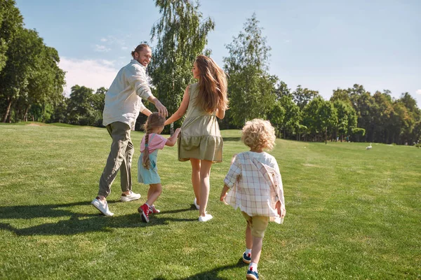 Bakåt syn på unga föräldrar som springer, leker på gräsplan med sina små barn, pojke och flicka på en sommardag — Stockfoto