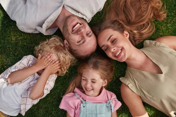 Vista superior de padres felices con niños pequeños y juguetones tumbados sobre hierba verde en el parque de verano —  Fotos de Stock
