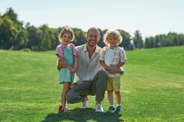 Gondoskodó apa mosolyog a kamerába, pózol együtt az aranyos kislányával és fiával, együtt állnak a fűpályán a nyári parkban. — Stock Fotó