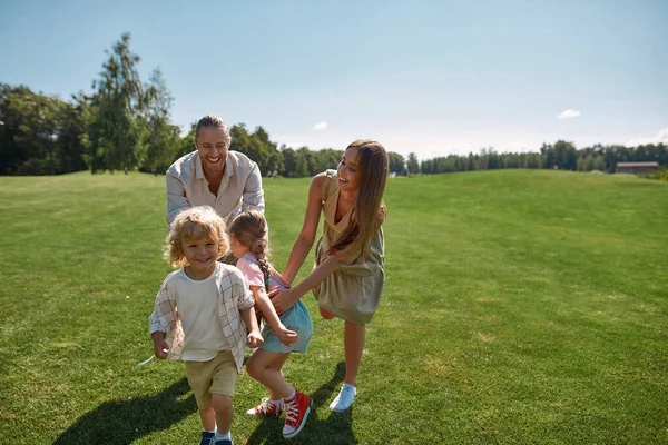 Yeşil Park 'ta bir yaz günü iki küçük çocuklu neşeli ebeveynler birlikte yakalamaca oynuyorlar. Mutlu aile boş zamanlarında eğleniyor. — Stok fotoğraf