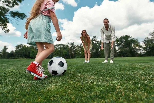 Gros plan de la petite fille qui donne un coup de pied au ballon pendant que les parents regardent. Jeune famille jouant au football sur le terrain d'herbe dans le parc un jour d'été — Photo