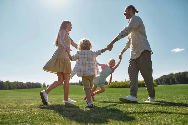 Happy family holding hands in circle, parents playing together with two little kids, boy and girl in the green park on a summer day — Stock fotografie