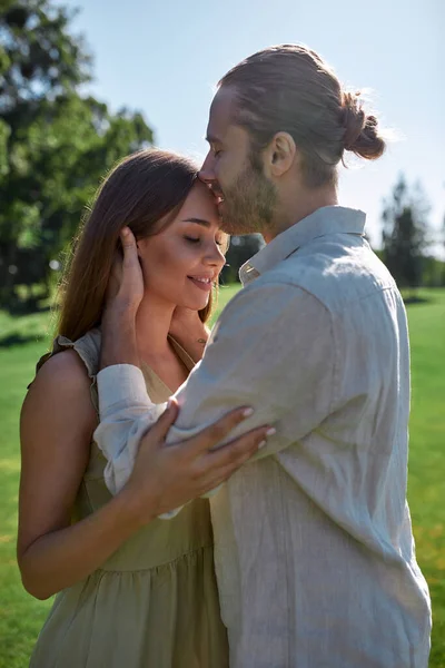Любящий муж целует улыбающуюся молодую жену в лоб, пока они позируют вместе на открытом воздухе в летнем парке — стоковое фото