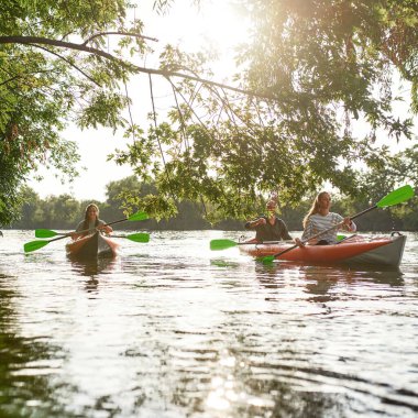İlkbahar ya da yaz aylarında ağaçlarla çevrili nehirde birlikte kayıkla gezen gençler açık havada vakit geçiriyorlar.