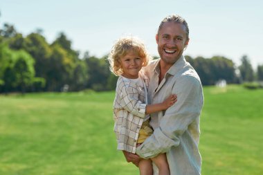 Heyecanlı genç baba kameraya gülüyor, oğluyla bir yaz günü parkta oynarken onu kucağına alıyor.