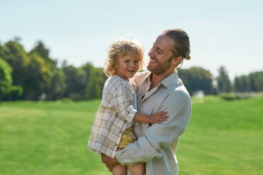 Sevgi dolu genç baba oğluna bakıyor, bir yaz günü çimen tarlasında oynarken ona sarılıyor.