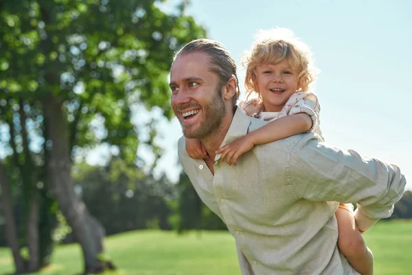 Boldog apa játszik a parkban, együtt mosolyog a kisfiával, aki a hátán ül. — Stock Fotó