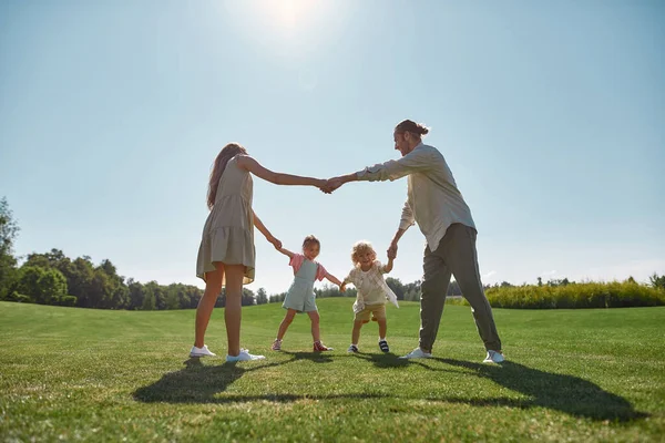 Šťastní mladí rodiče tráví čas spolu, drží se za ruce v kruhu se svými dvěma malými dětmi, chlapec a dívka v zeleném parku v letní den — Stock fotografie