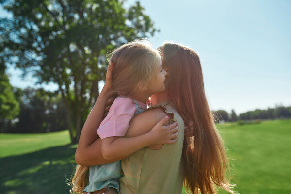 Loving mother playing with her cute little daughter, holding her while spending time together in summer park