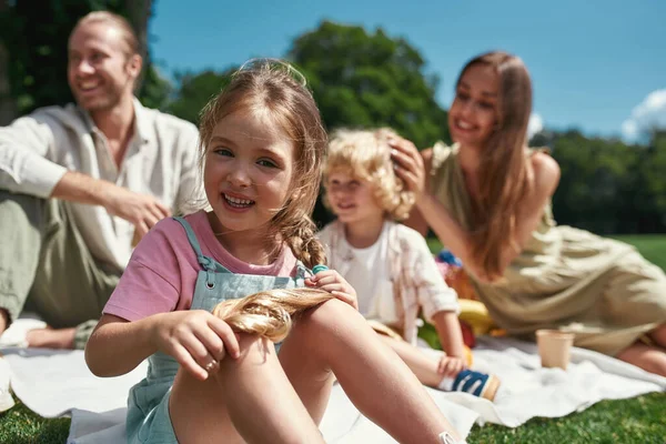 Adorable niña sonriendo a la cámara mientras se relaja con los padres y el hermano al aire libre, haciendo un picnic en la naturaleza en un día de verano —  Fotos de Stock