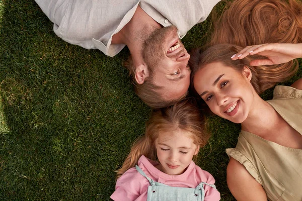 Bovenaanzicht van gelukkige jonge ouders met kleine dochter liggend op groen gras in zomerpark — Stockfoto