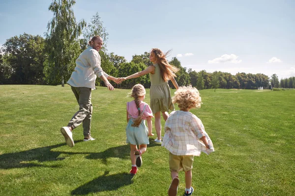 Padres alegres tomados de la mano, huyendo de sus hijos pequeños, niños y niñas, divirtiéndose juntos en el parque verde en un día de verano — Foto de Stock