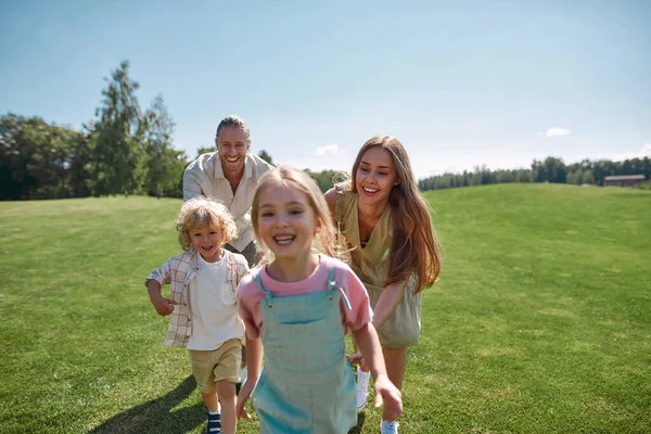 Jóvenes padres alegres se divierten juntos con dos niños pequeños, niño y niña en el parque verde en un día de verano —  Fotos de Stock