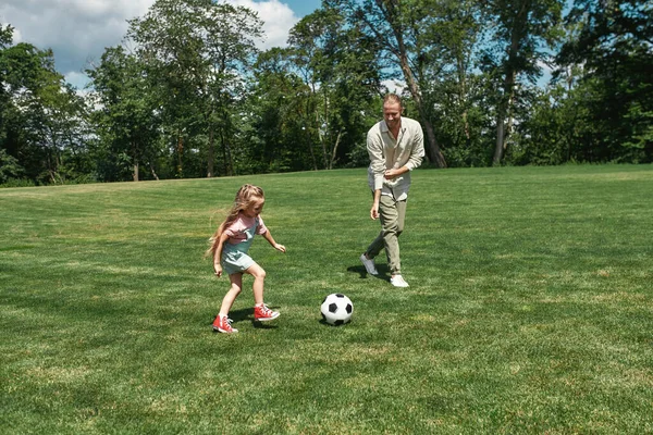 Jeune père actif jouant au football avec sa petite fille sur le terrain d'herbe dans le parc un jour d'été — Photo