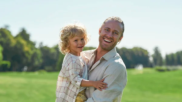 Glada unga pappa skrattar åt kameran, håller sin lilla son medan du leker med honom i parken på en sommardag — Stockfoto