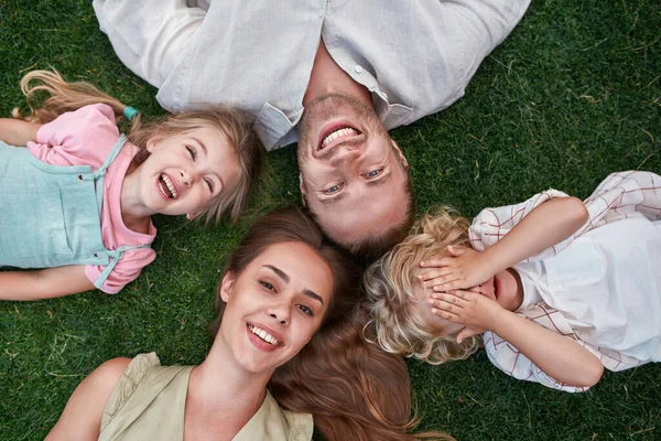 Retrato de una alegre familia joven con una linda niña y un niño riéndose de la cámara mientras se relajan, tumbados juntos sobre hierba verde en el parque de verano —  Fotos de Stock