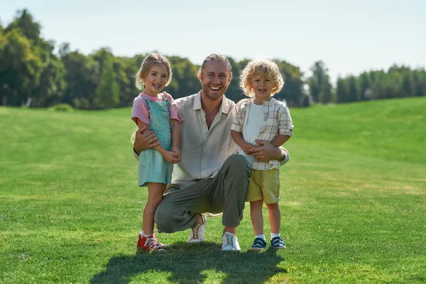Full längd skott av glad far ler mot kameran, poserar med sin söta lilla dotter och son, står tillsammans på gräsplan i sommarparken — Stockfoto