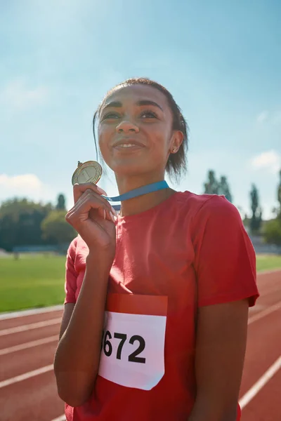Porträtt av ung kvinnlig idrottsman mästare ler bort och håller hennes guldmedalj när du står på banan — Stockfoto