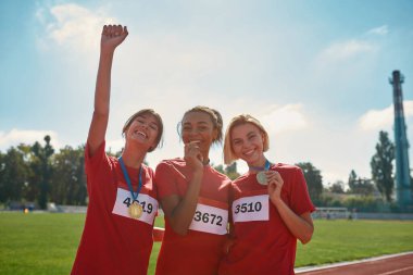 Altın madalyaları olan neşeli genç bayan sporcular yarış pistinde birlikte duruyorlar.