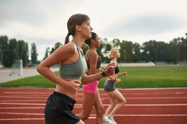 Vista lateral del grupo de tres jóvenes corredores profesionales corriendo juntos en el campo de atletismo en el estadio — Foto de Stock