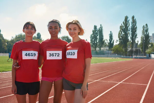 Grupp av unga professionella kvinnliga löpare bär t-shirts med nummer tittar på kameran när de står tillsammans, redo att starta loppet — Stockfoto