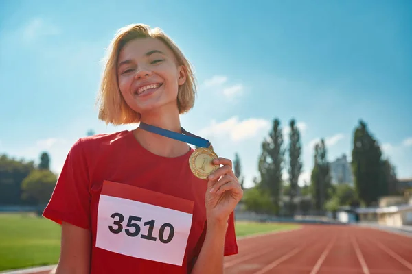 Glada unga kvinnliga idrottare ler mot kameran och visar sin guldmedalj när hon står på tävlingsbanan — Stockfoto