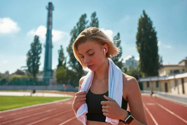 Porträtt av attraktiv sportig ung kvinna med handduk runt halsen promenader längs banan på stadion på en solig dag — Stockfoto