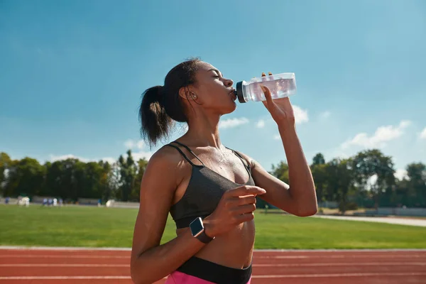 Professionell ung kvinnlig löpare dricksvatten efter träning, står på banan på stadion på en solig dag — Stockfoto