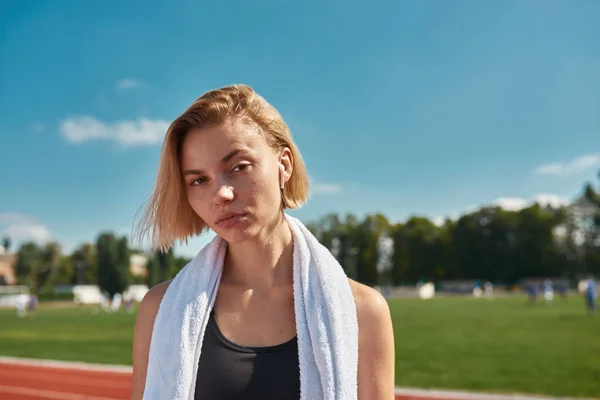 Güneşli bir günde stadyumdaki pistte poz verirken boynunda havluyla kameraya bakan ciddi sportif genç bir kadının portresini yakından çek. — Stok fotoğraf