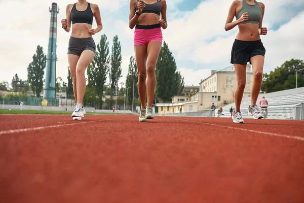 Cropped shot of three professional young female runners in sportswear running together on track field at the stadium