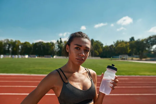 Porträtt av svettiga professionella unga kvinnliga löpare tittar på kameran, står med flaska vatten på banan på stadion på en solig dag — Stockfoto