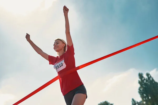 Låg vinkel syn på glad ung kvinnlig löpare med armarna upp når mållinjen på banan under maraton utomhus — Stockfoto