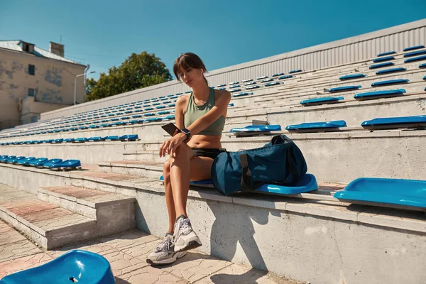 건강 한 젊은 여성 이 스마트폰을 사용하는 장면, 운동 후 휴식을 취하는 동안 소셜 미디어에서 식사를 하는 장면, 야외 경기장 의자에 앉아 있는 모습 — 스톡 사진