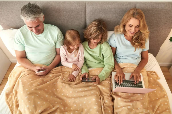 Szczęśliwa rodzina z dziećmi pobyt w łóżku korzystania urządzeń — Zdjęcie stockowe