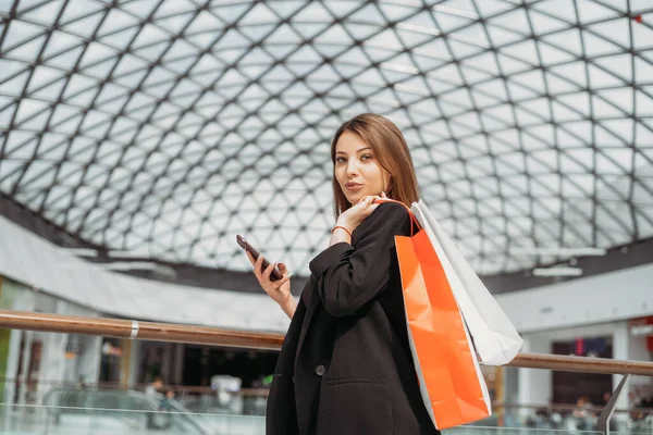 Schönes dunkelhaariges Mädchen mit Einkaufstaschen und Handy auf verschwommenem Hintergrund — Stockfoto