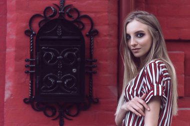 Uzun saçlı genç model kırmızı tuğlalı duvarda poz veriyor.