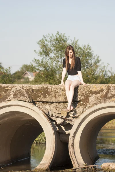 Belle fille aux cheveux longs et raides posant et jouant sur un pont en béton sur une petite rivière — Photo