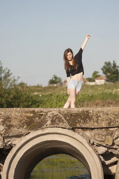 Mooi meisje met lange, rechte haren poseren en spelen op een betonnen brug over een kleine rivier — Stockfoto