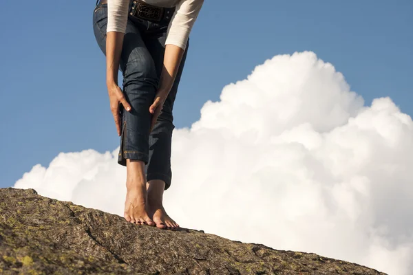 Pernas de mulher, pés e mãos com jeans, sobre uma rocha, com fundo natural e nuvens brancas fofas — Fotografia de Stock