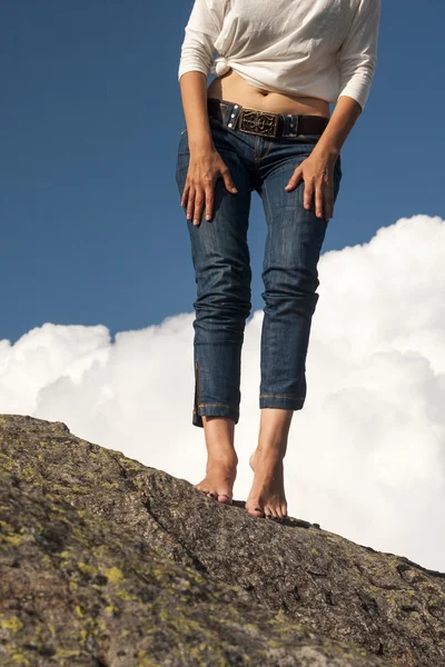 Pernas de mulher, pés e mãos com jeans, sobre uma rocha, com fundo natural e nuvens brancas fofas — Fotografia de Stock