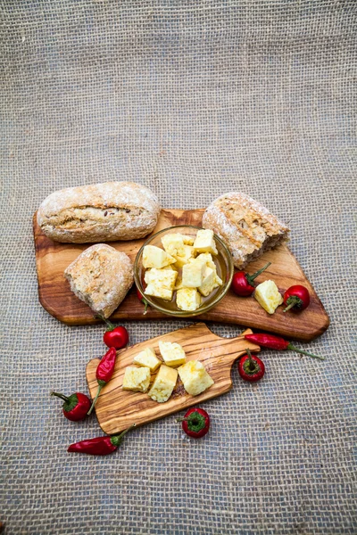 Composizione con legno di ulivo, pezzi di formaggio in olio d'oliva, pane e peperoncino con texture di iuta sullo sfondo — Foto Stock