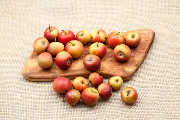 Pequeñas manzanas rojas y tabla de cortar de cocina con textura de arpillera en el fondo —  Fotos de Stock