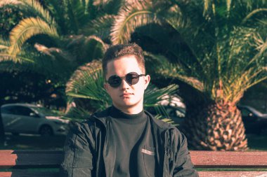 Genç bir adam, palmiye ağaçlarının arkasında bir bankta oturuyor..